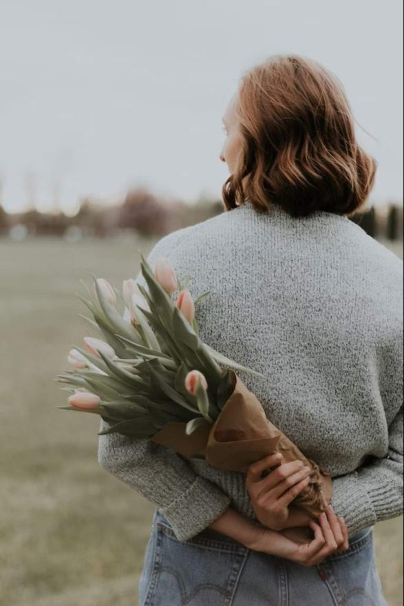 Žena drži cvijeće iza leđa i gleda u daljinu - Koje cvijeće pokloniti majci? 